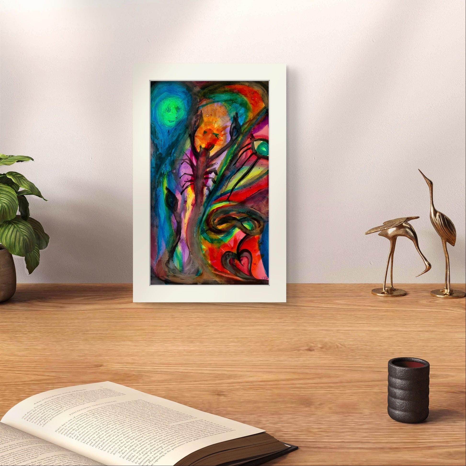 Watercolor print on wooden desk with a book in front of white wall.