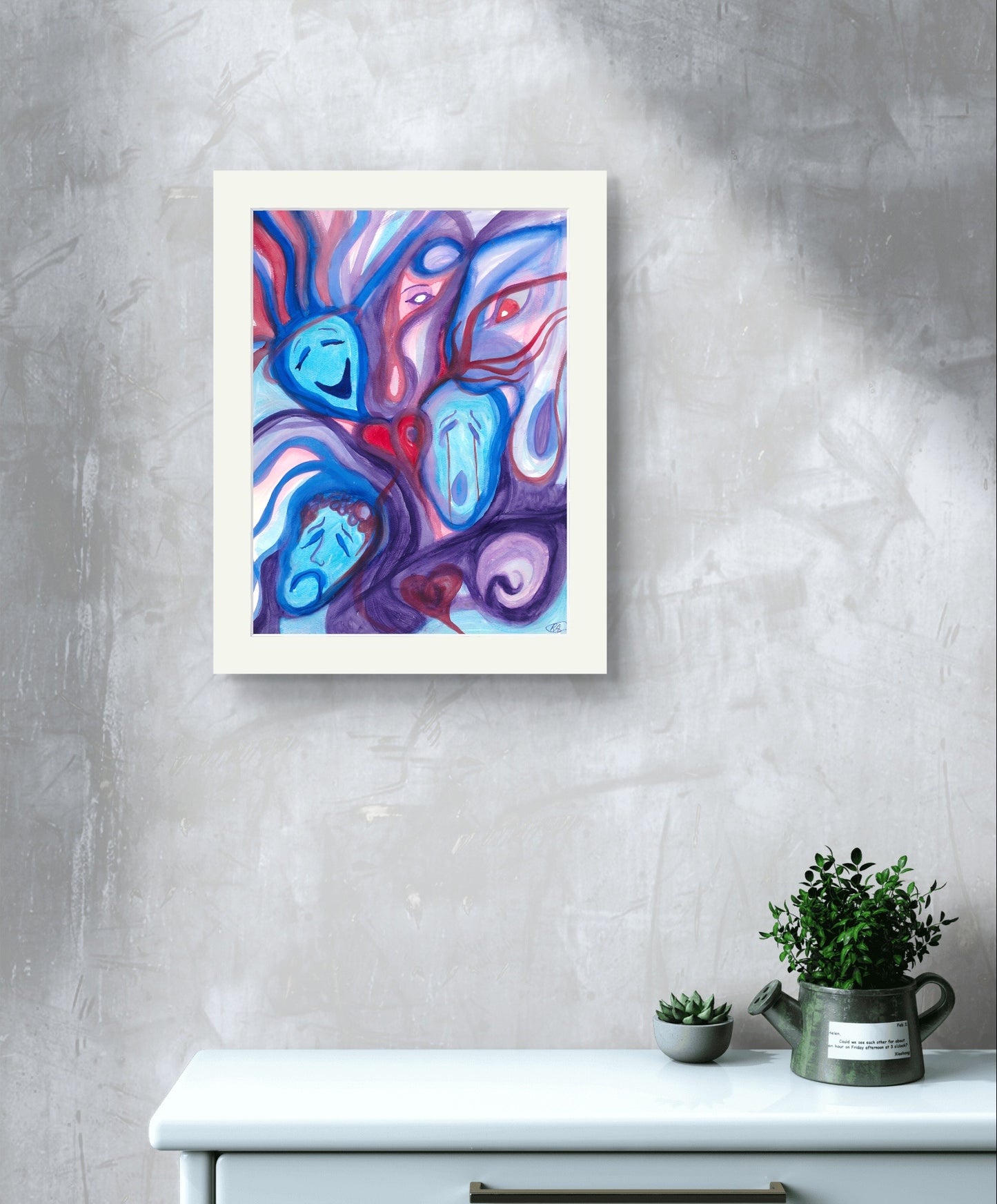 White bordered version of faces painting on grey wall above white table and plant.